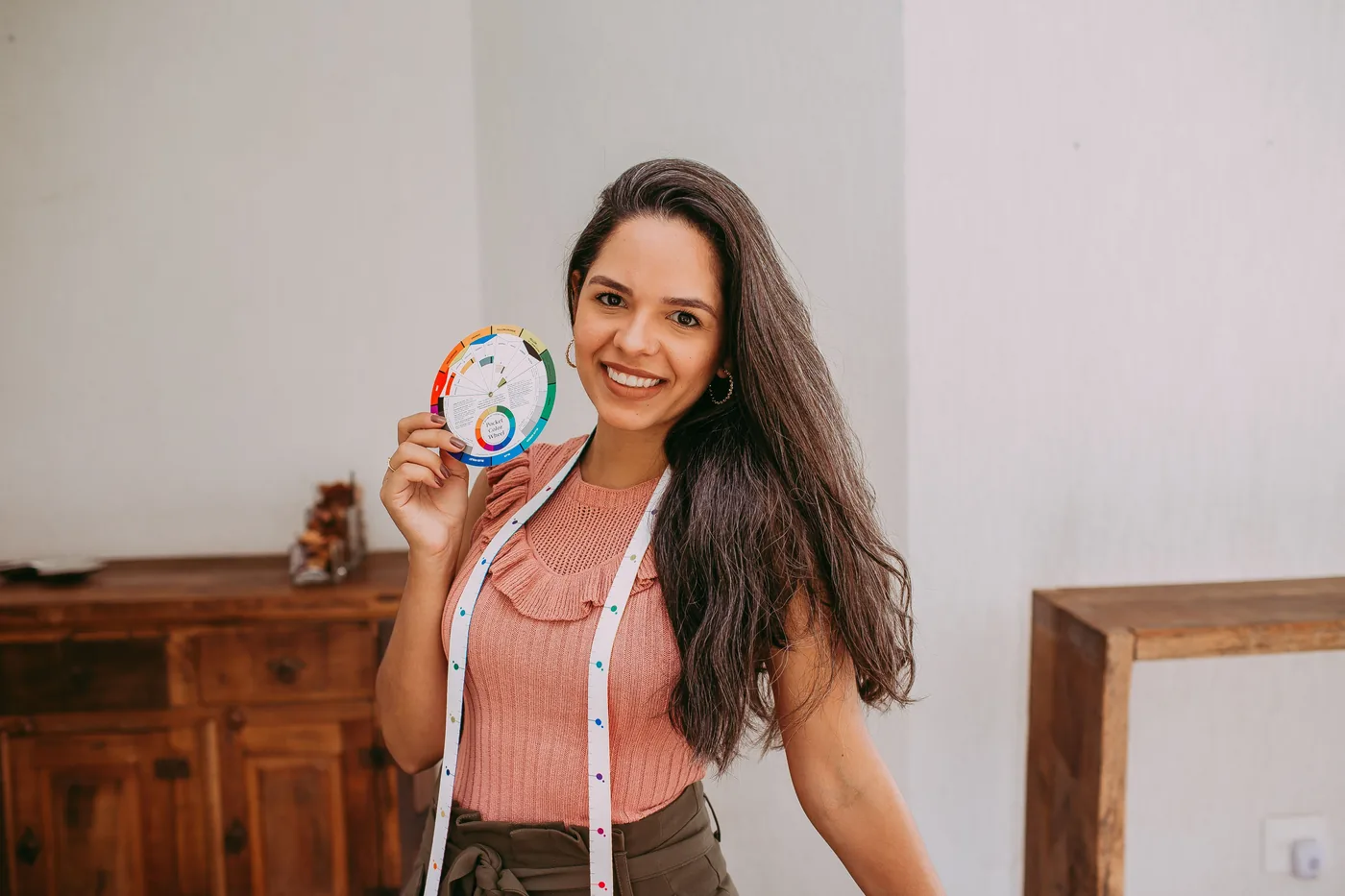 Empreendedora sorrindo no seu ateliê, com fita métrica no pescoço e uma roda de cores na mão