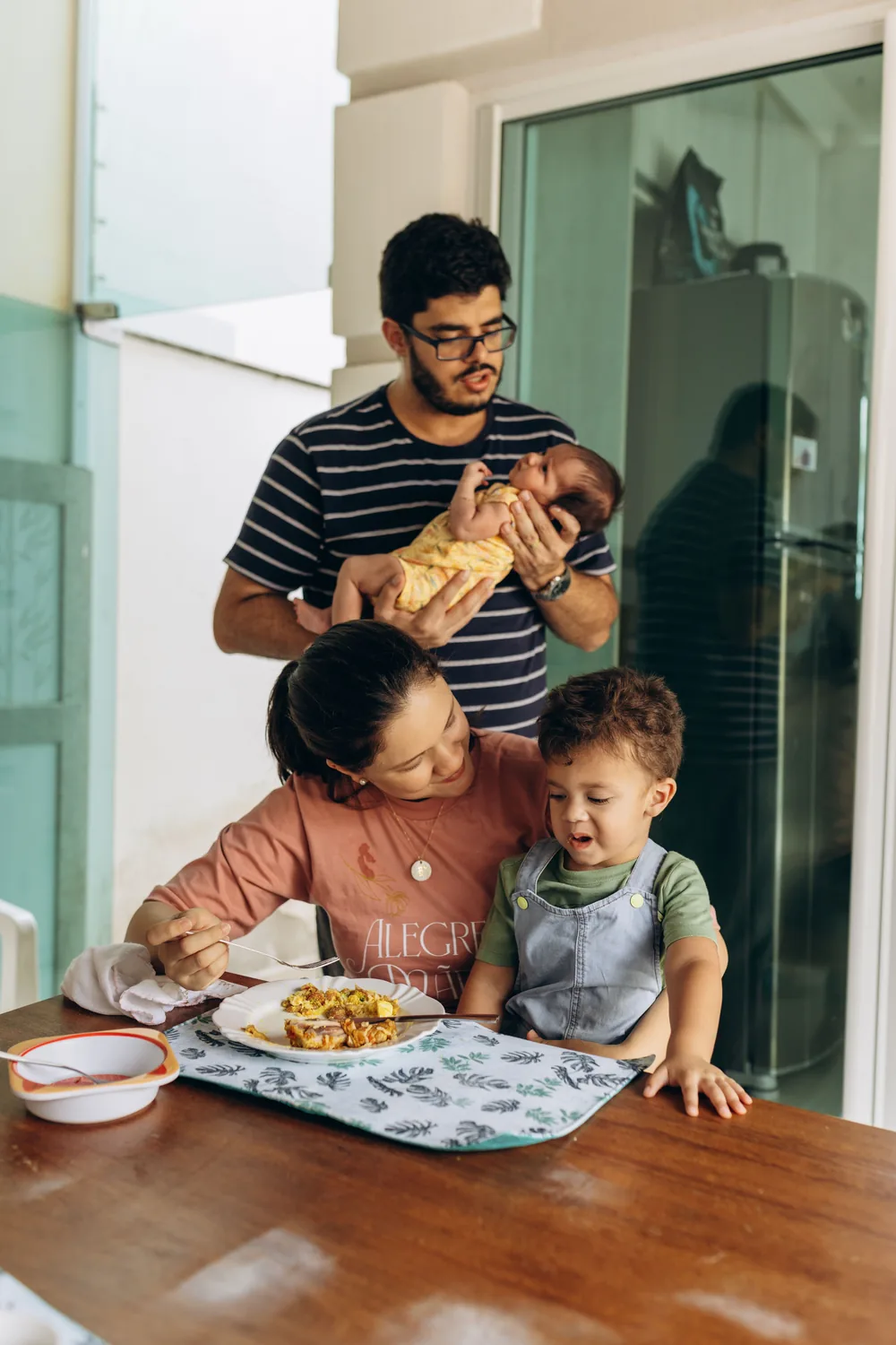 Família à mesa em casa, a mãe com o filho no colo e o pai com o bebê