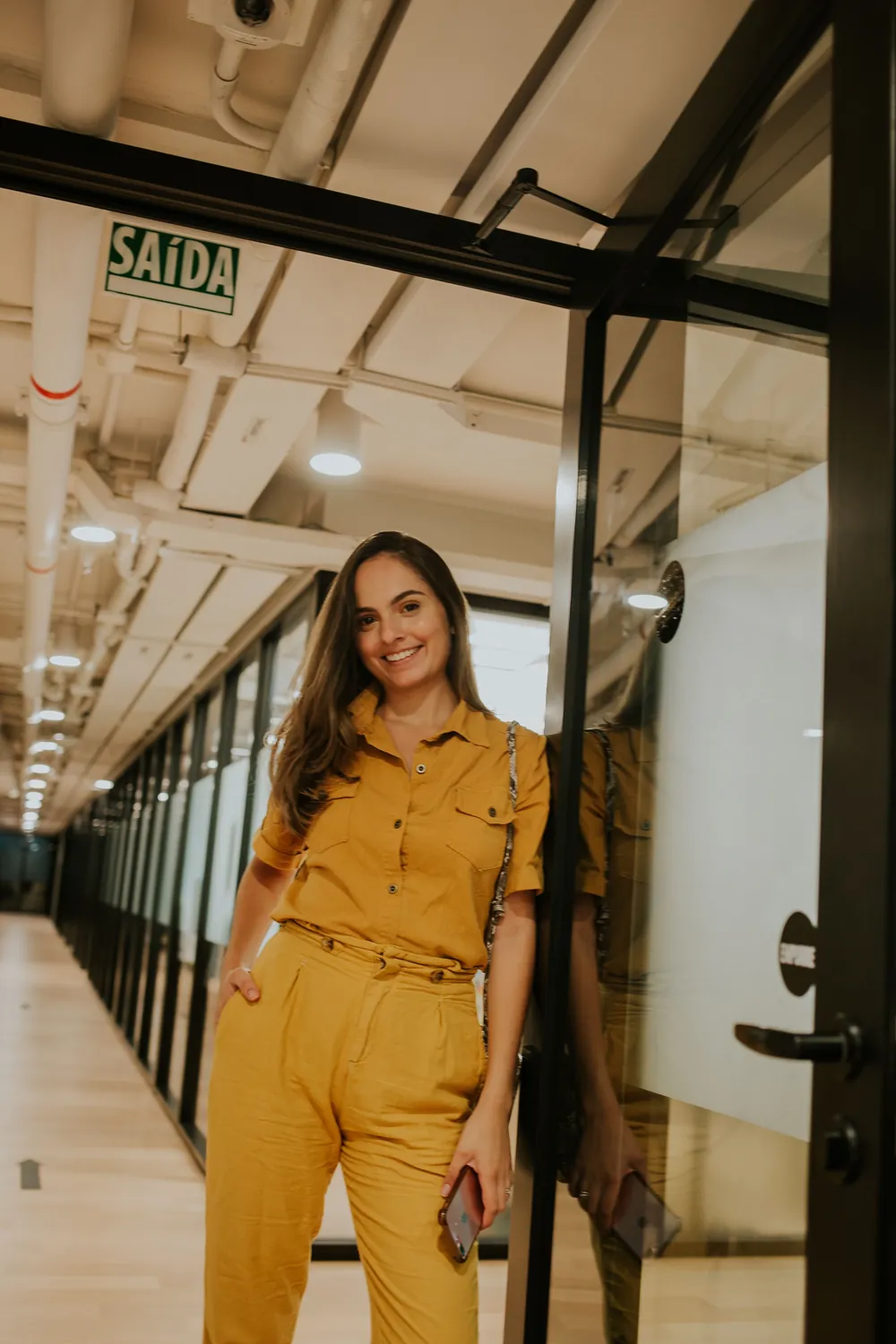Mulher sorrindo com o celular na mão, no corredor de um escritório