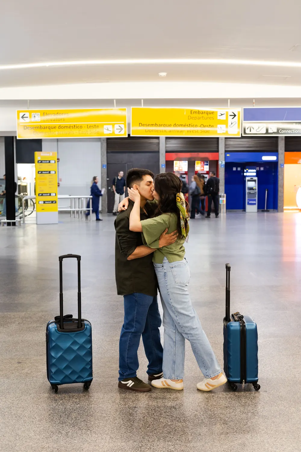 Casal se beijando com as malas no saguão do aeroporto, perto da placa de embarque